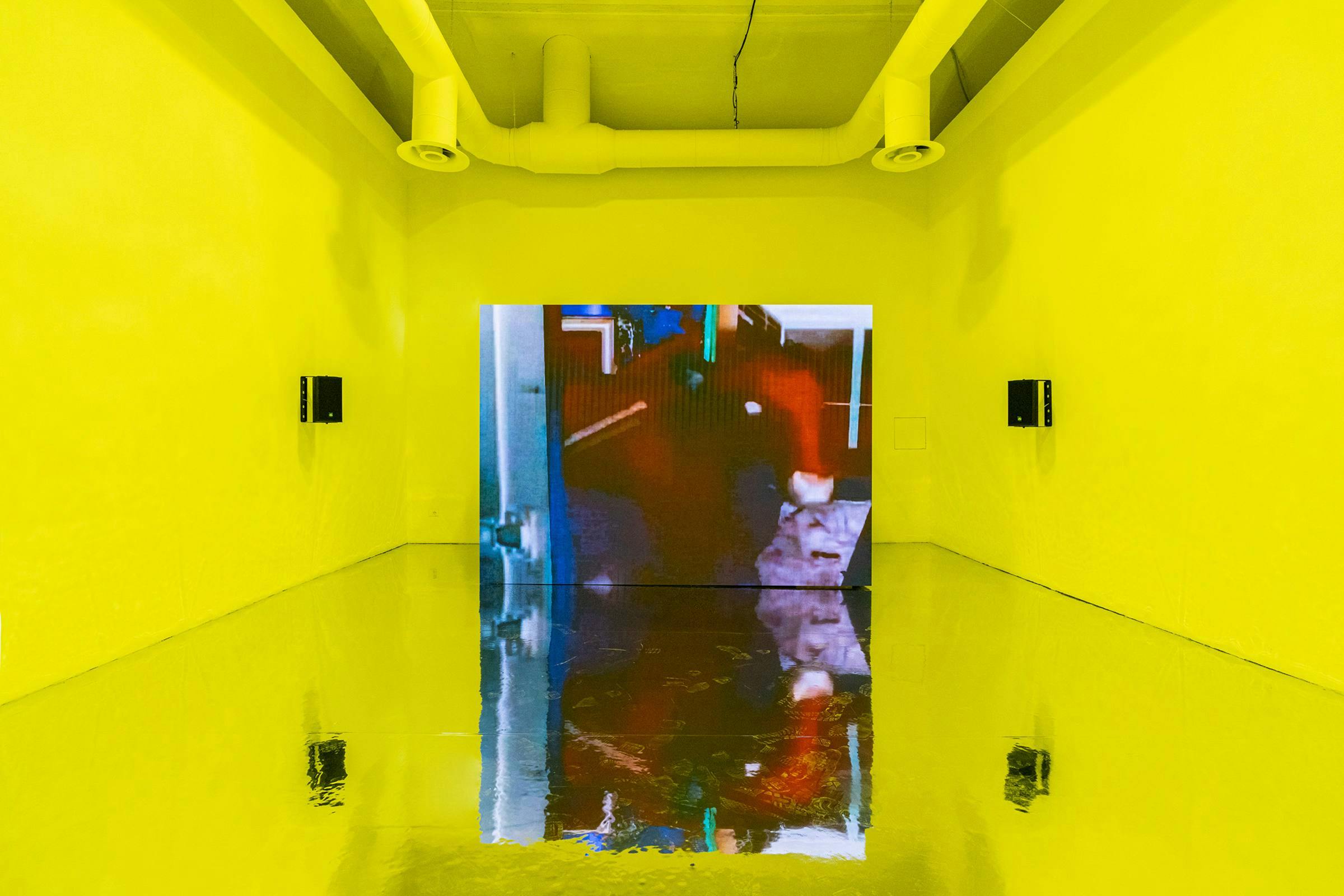 a video artwork installed in an art gallery. The walls are painted yellow and the floor is made from a highly reflective material.