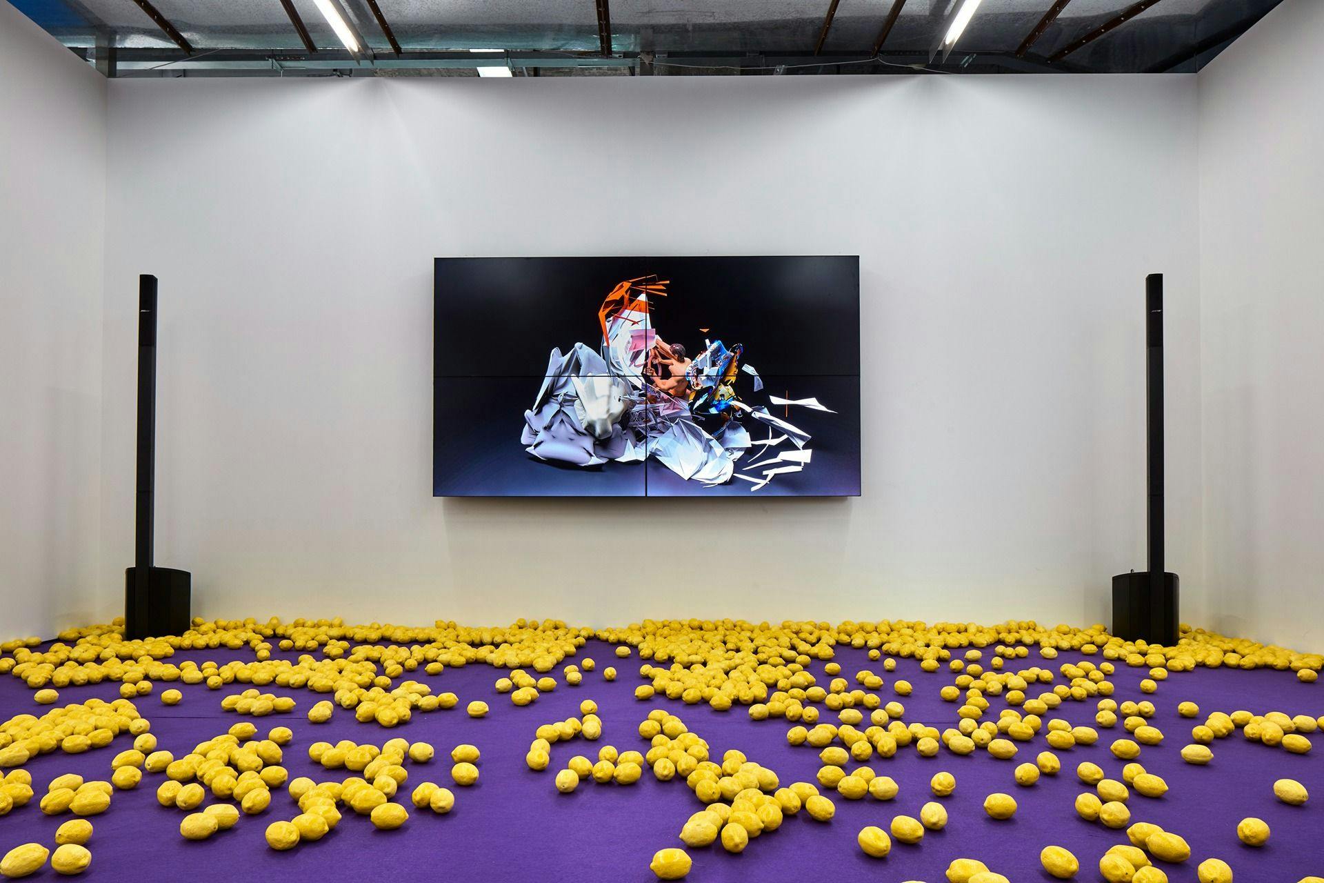 Installation view of Installation view, Samson Young, The highway is like a lion's mouth, 6 November - 23 December 2018, Edouard Malingue Gallery, Shanghai. A tv screen with a still of the video is hanging on a white wall. There are 2 freestanding speakers either side. The floor is carpeted in purple carpet and there are yellow objects scattered across the floor. It is difficult to discern what these objects are.