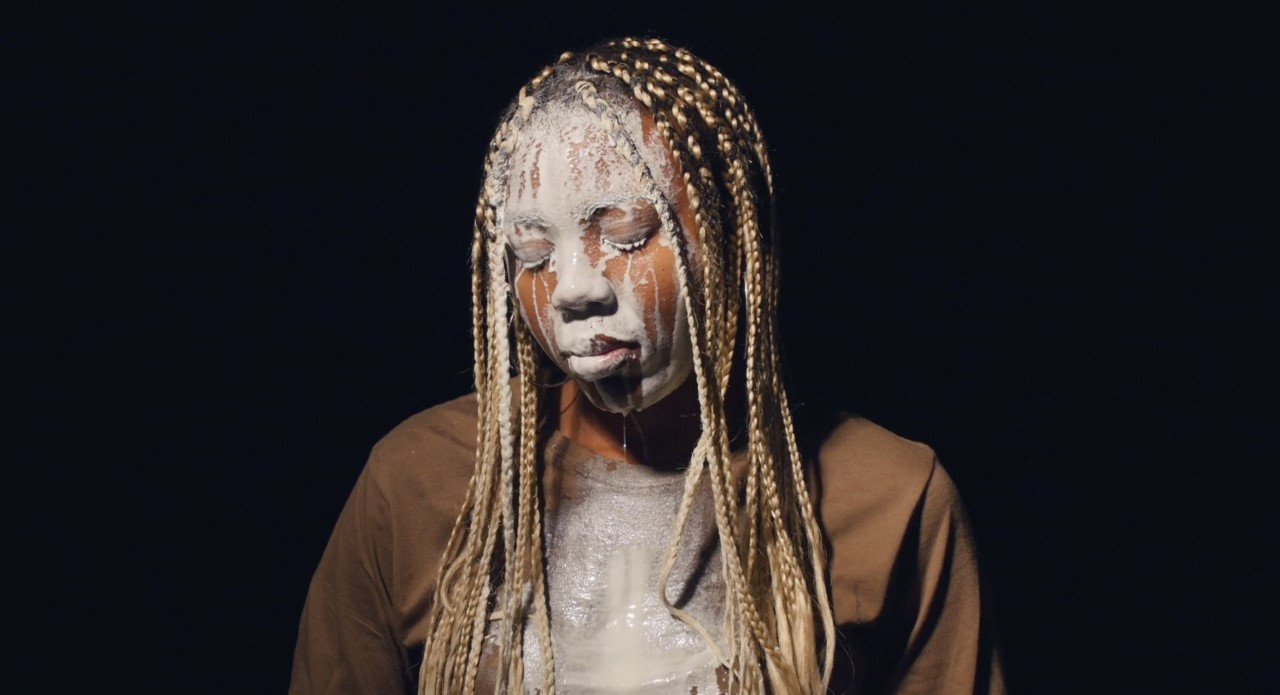 A black women with long hair and wearing a brown jacket face is covered in milk. The background is black.